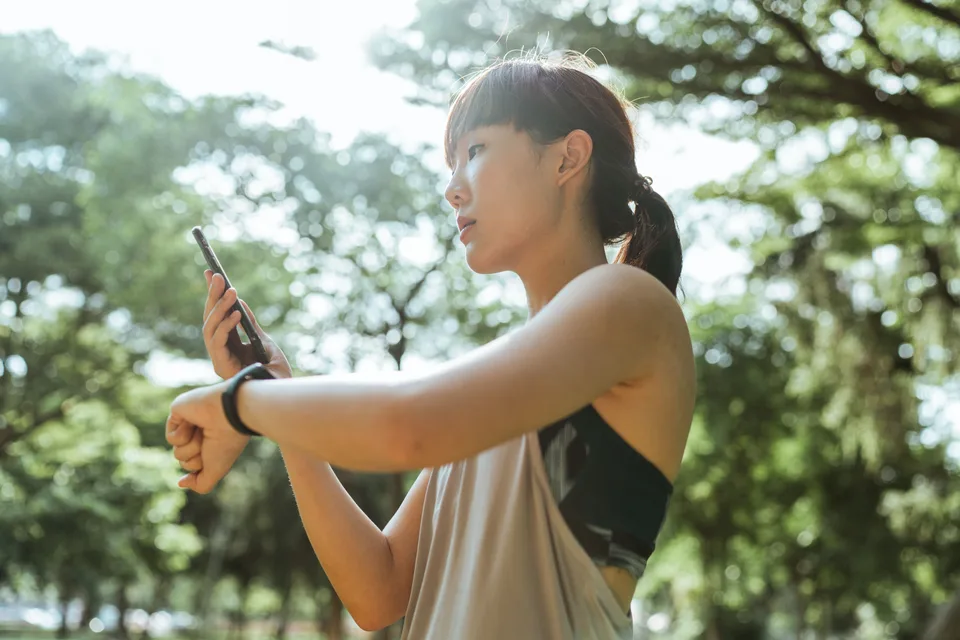 A young Asian woman using fitness devices and her phone while exercising outdoors in a park.