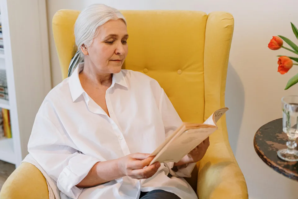 An elderly woman sitting comfortably on a couch reading a book in a warm domestic setting.