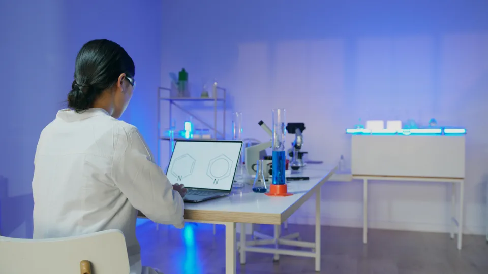 A scientist working at a laptop displaying a complex chemical formula on the screen.