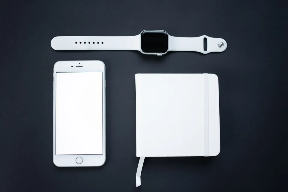 white smartwatch with a sport band laid above a smartphone and a notebook on a dark surface