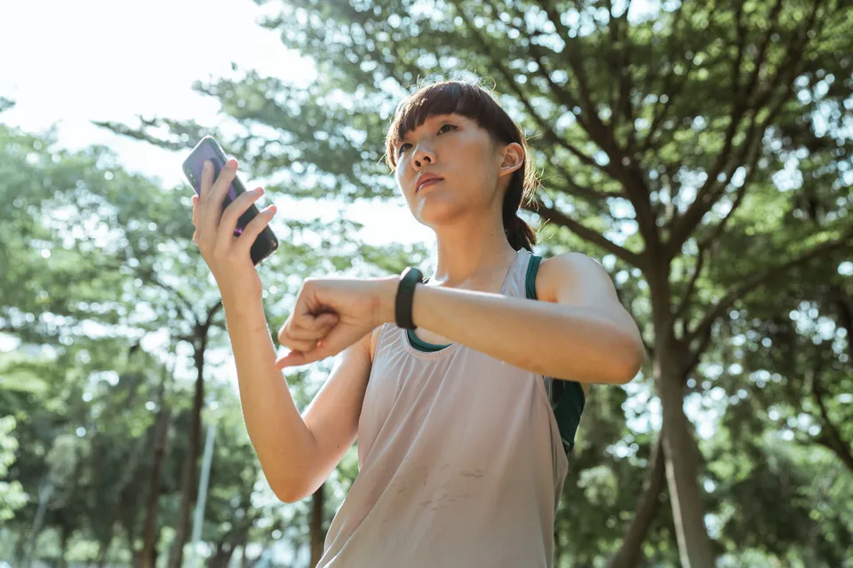 woman in a park holding a smartphone while checking a fitness band on her wrist