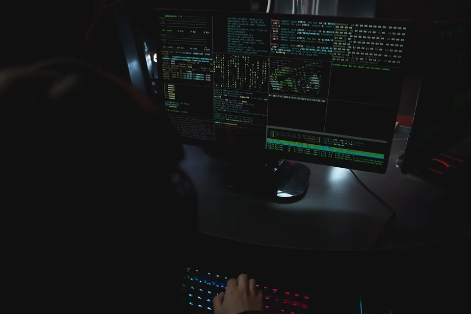 person typing on a backlit keyboard in front of a monitor full of terminal windows in a dark room
