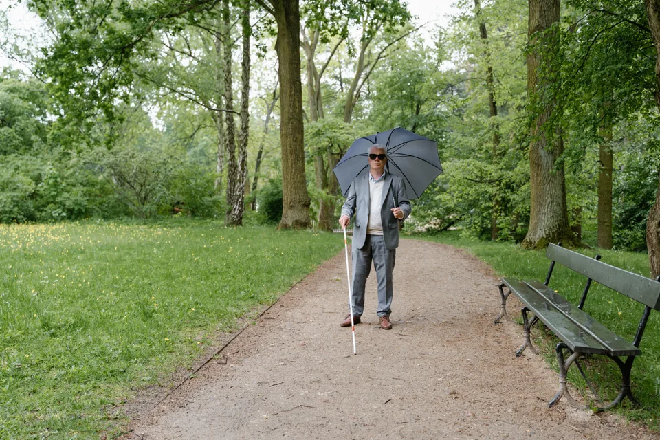 An elderly man standing on a pathway holding an umbrella and a walking stick.