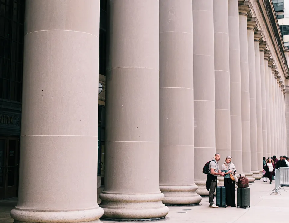 two travelers with suitcases standing beside a long row of massive stone columns outside a classical building