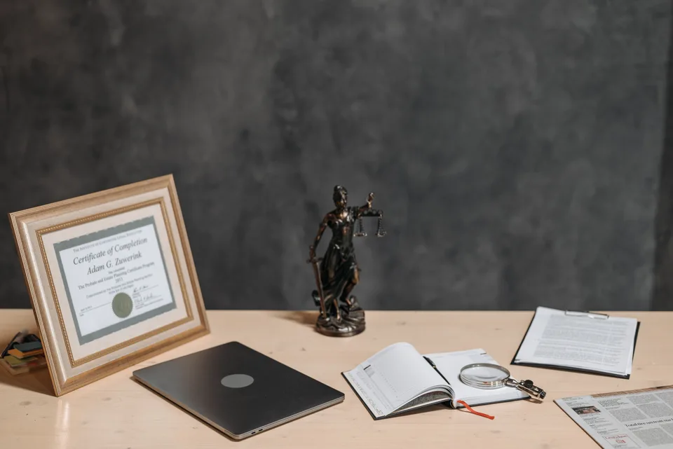 bronze scales-of-justice statuette on a wooden desk beside a closed laptop, an open planner with a magnifying glass and a clipboard of papers