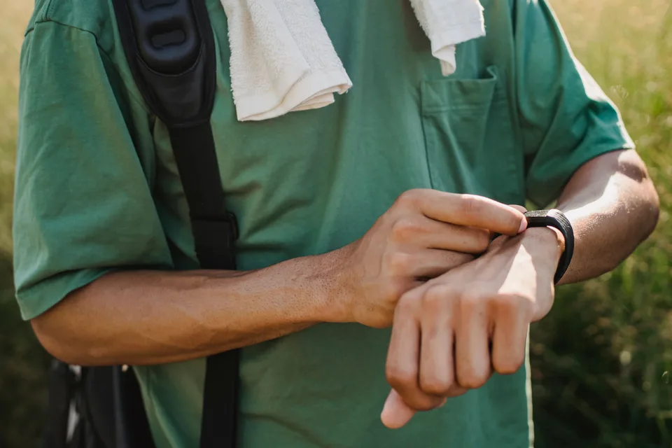 man in a green t-shirt with a backpack strap and towel pressing a button on a black sports watch outdoors