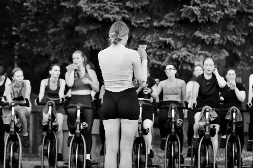 An instructor leads a group spin class with women on stationary bicycles in a studio.