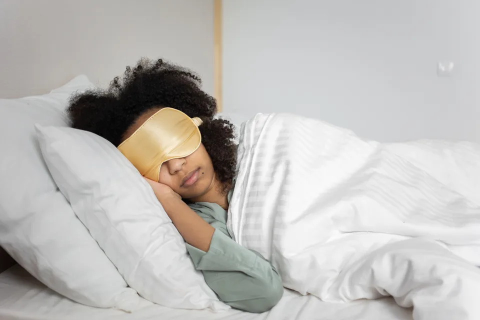 woman wearing a satin sleep mask asleep in a bed with white sheets