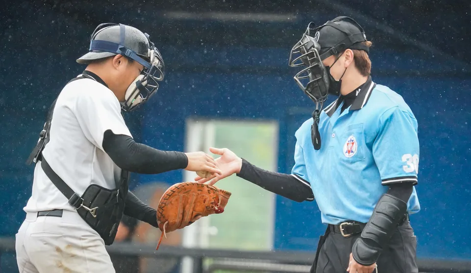 baseball catcher in mask and chest protector handing a ball to a home plate umpire in a light blue shirt