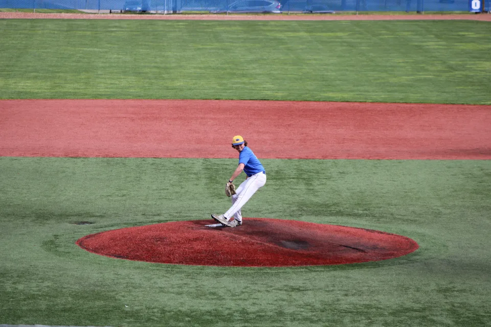 baseball pitcher in a blue jersey delivering a pitch from a red mound on a green field