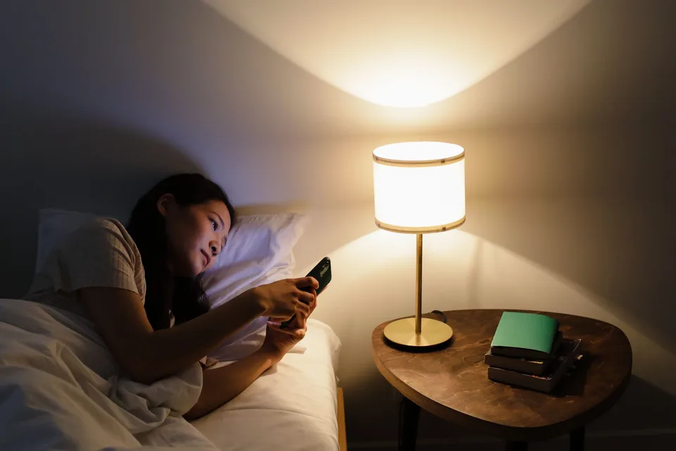 Woman lying in bed at night looking at her smartphone beside a lit bedside lamp