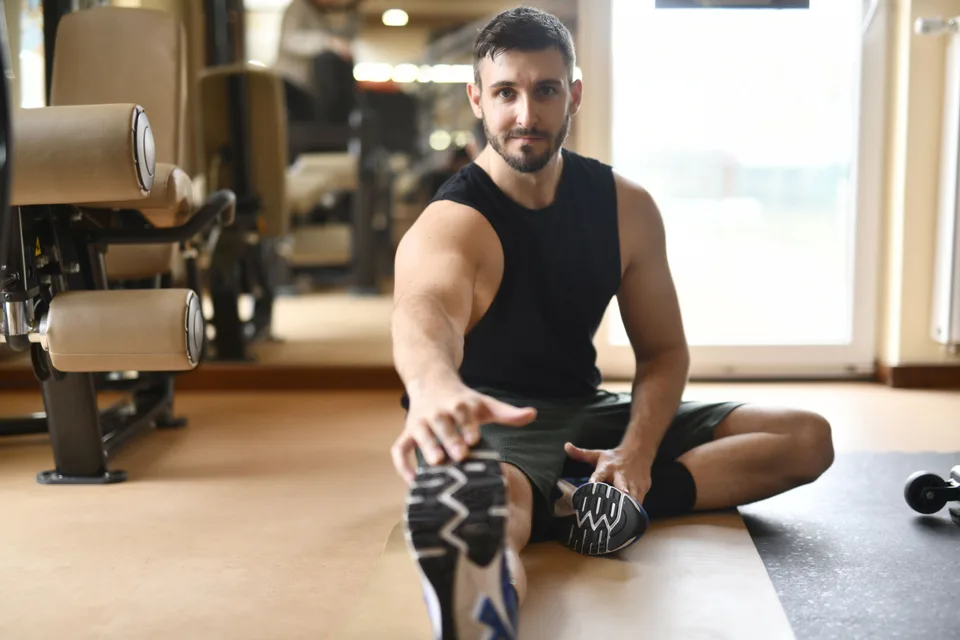 A man in a black tank top stretching in a gym setting as part of a post-workout routine.