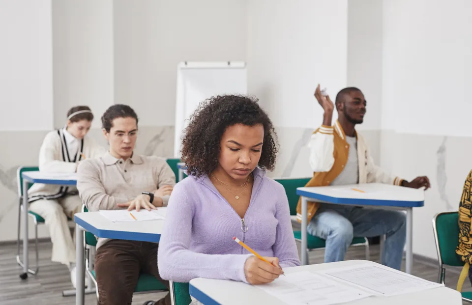 students seated at classroom desks writing answers on paper test sheets with pencils