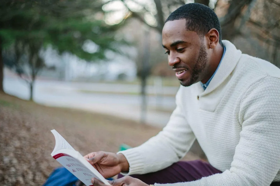 A man in a sweater reading a book in a relaxed indoor setting.