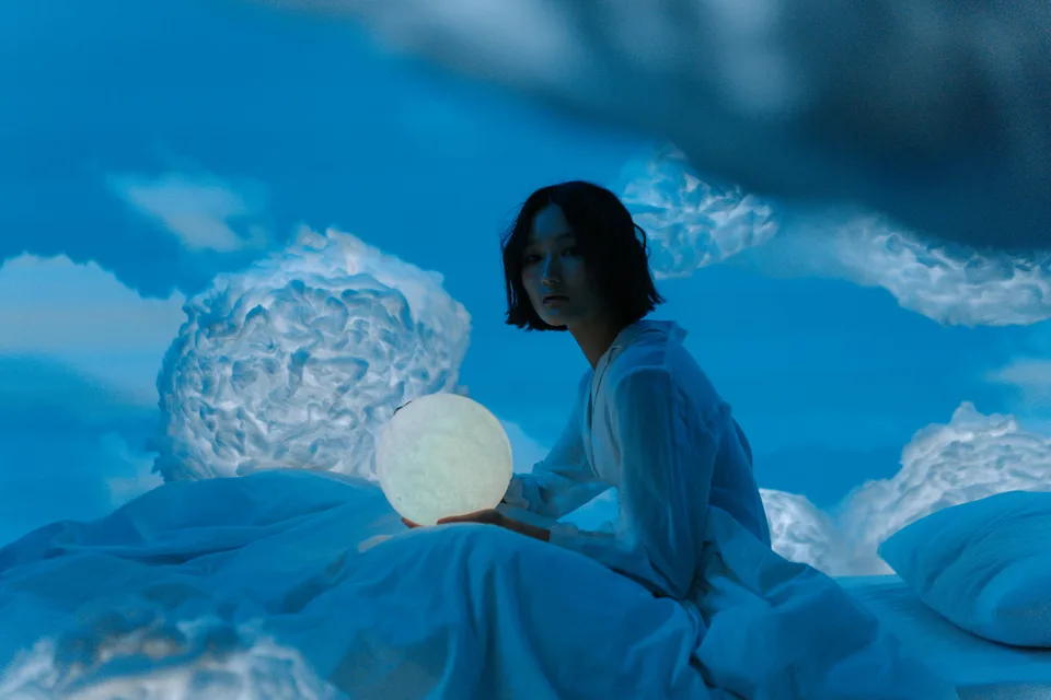 A person sitting on a bed in a calm bedroom setting holding a moon lamp.