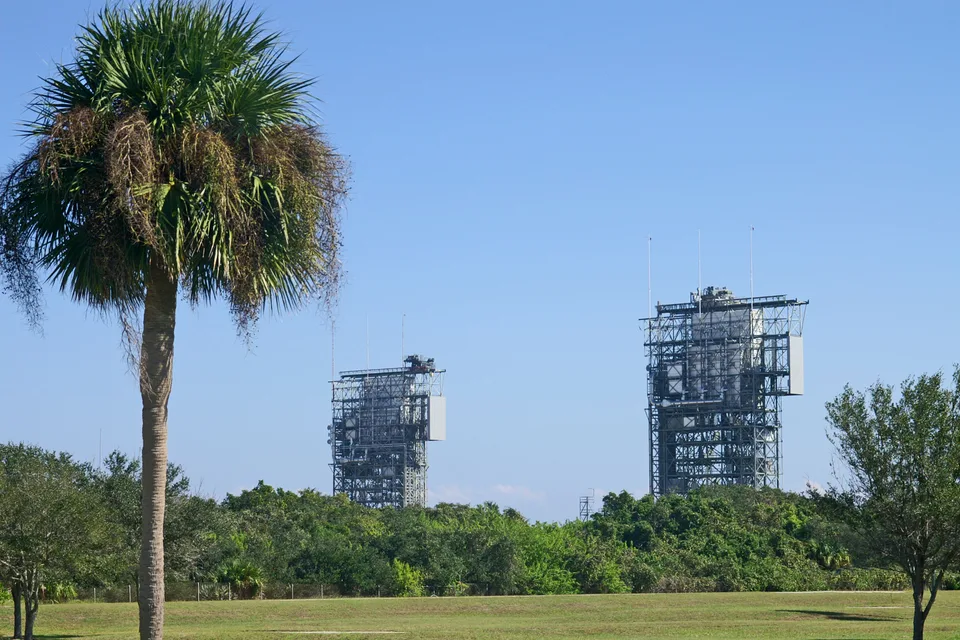 Two steel rocket launch towers rising above green trees under a clear blue sky, with a palm tree in the foreground