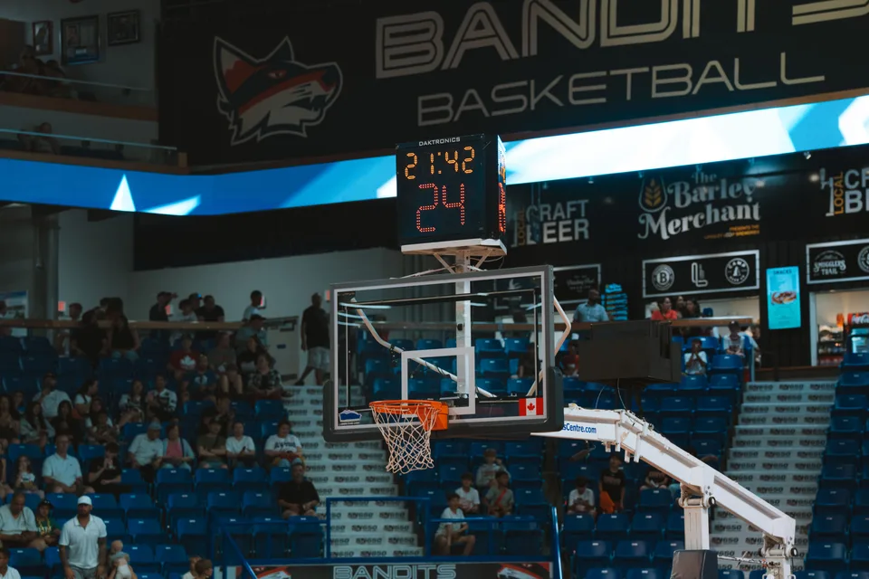 An indoor basketball court with a scoreboard and audience visible in the stands.