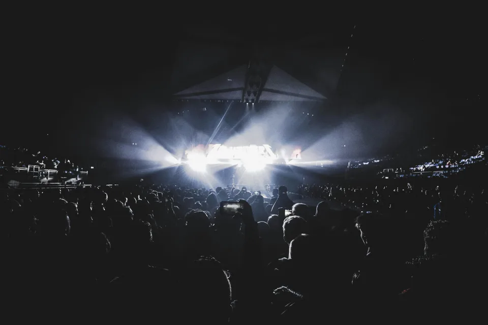 A large crowd watching a live event inside a dark stadium with bright stage lighting.